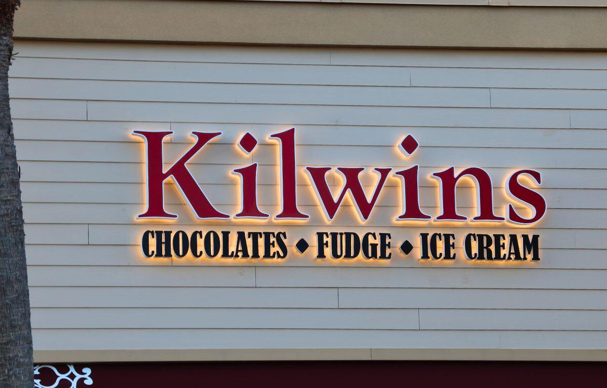 A light up sign on a beige building reads, "Kilwins. Chocolate. Fudge. Ice Cream." A palm tree stands to the left of it.