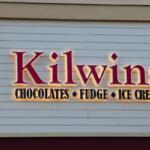 A light up sign on a beige building reads, "Kilwins. Chocolate. Fudge. Ice Cream." A palm tree stands to the left of it.
