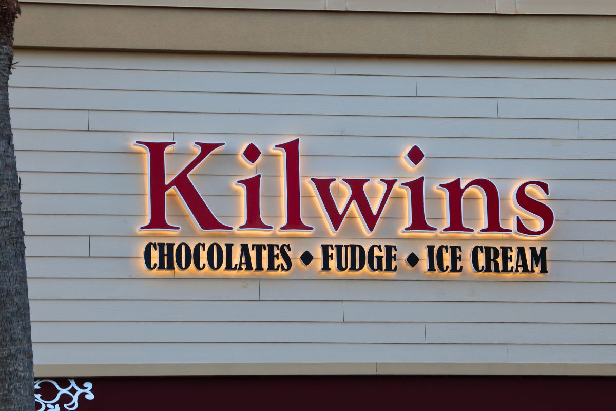 A light up sign on a beige building reads, "Kilwins. Chocolate. Fudge. Ice Cream." A palm tree stands to the left of it.