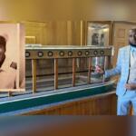 A portrait of a man in a uniform on the left, with a model of a glass-bottom boat, with a man in a blue suit standing to the right, with a white wall in the background, with an open door on the far right.