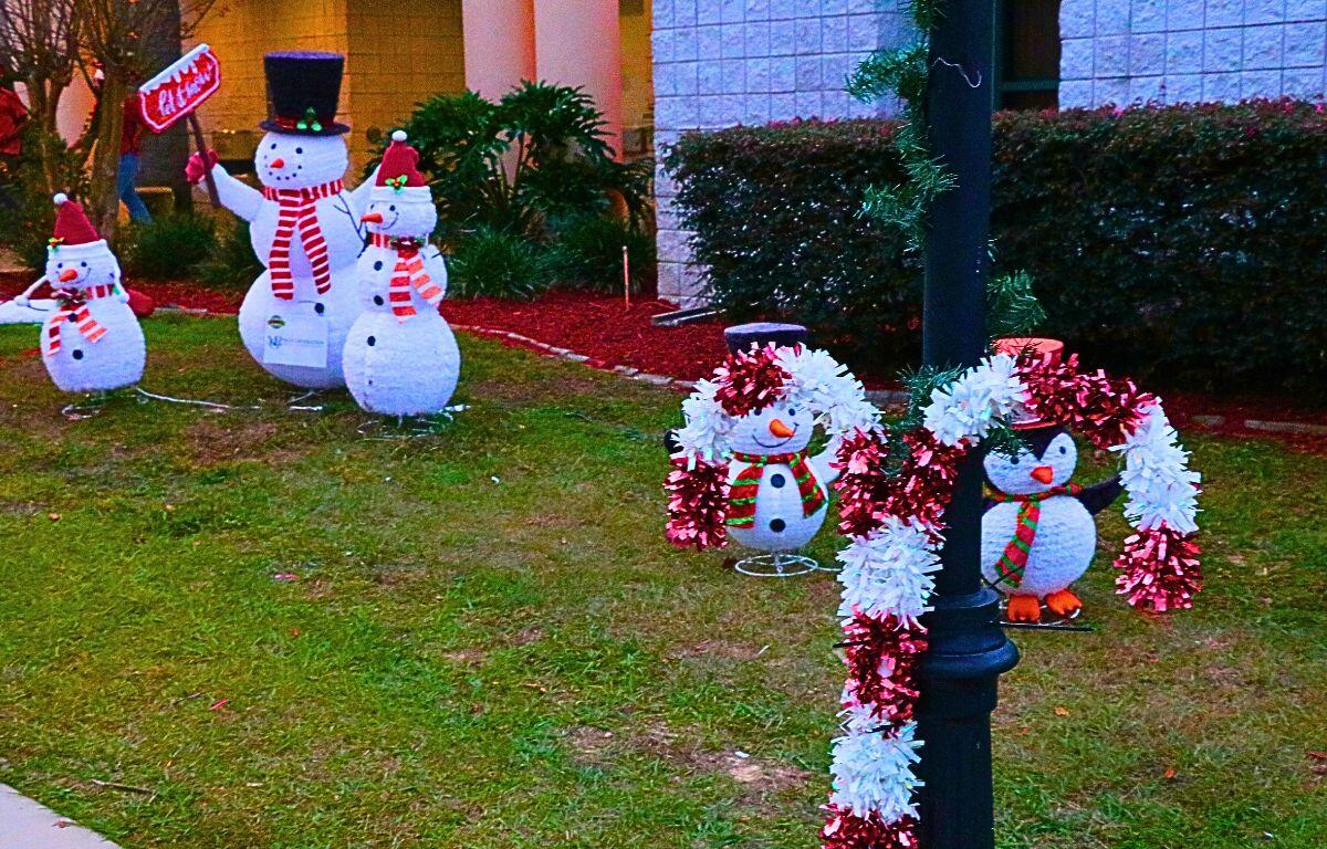 A candy cane decoration stands against a light pole with snowmen and a brick building in the background.