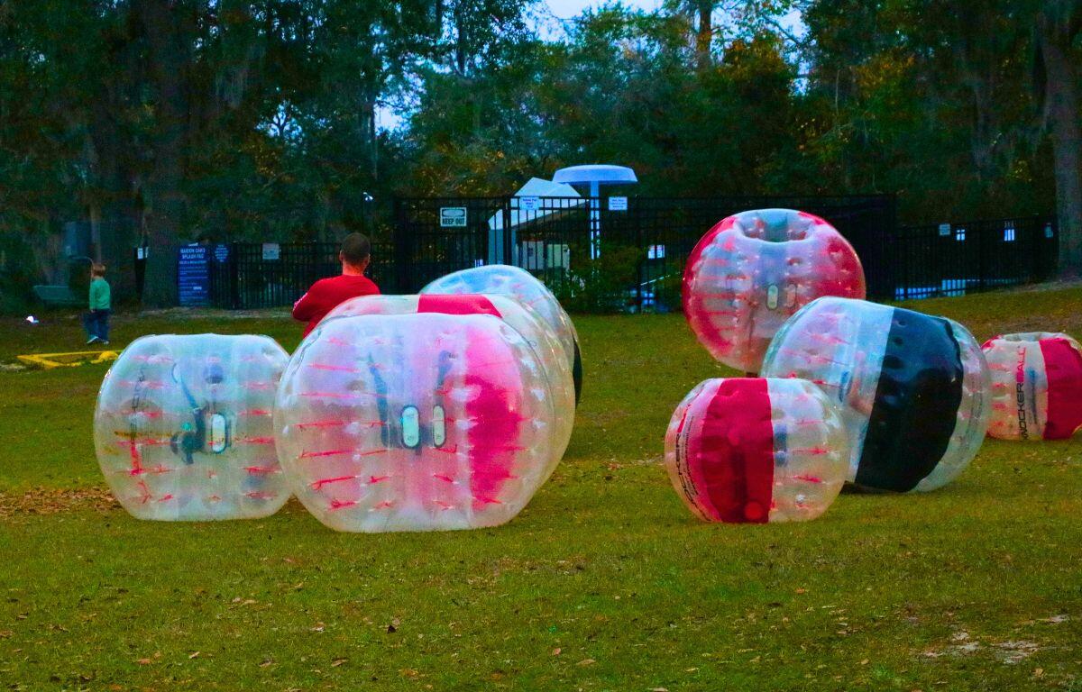 Large human hamster balls are scattered in an open field with kids bumping into each other inside.
