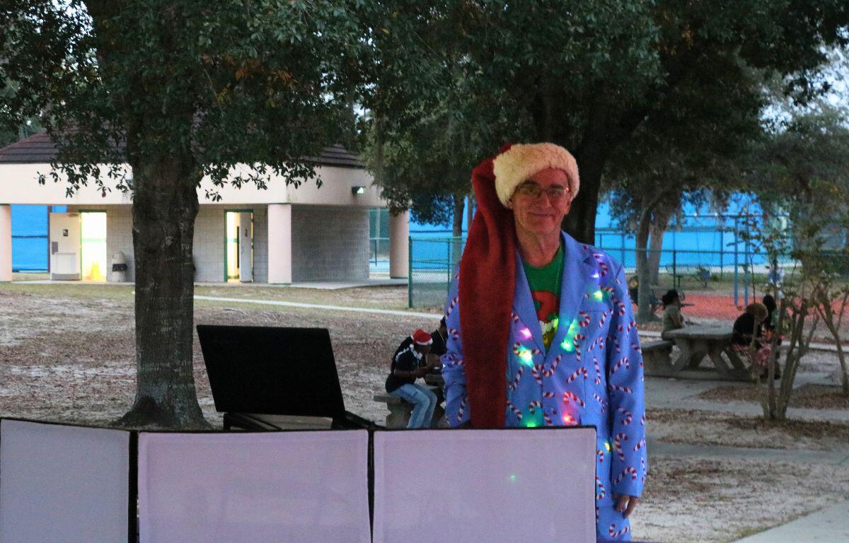 A man stands behind a computer playing music in a candy cane suit with a grinch shit underneath and Santa hat.