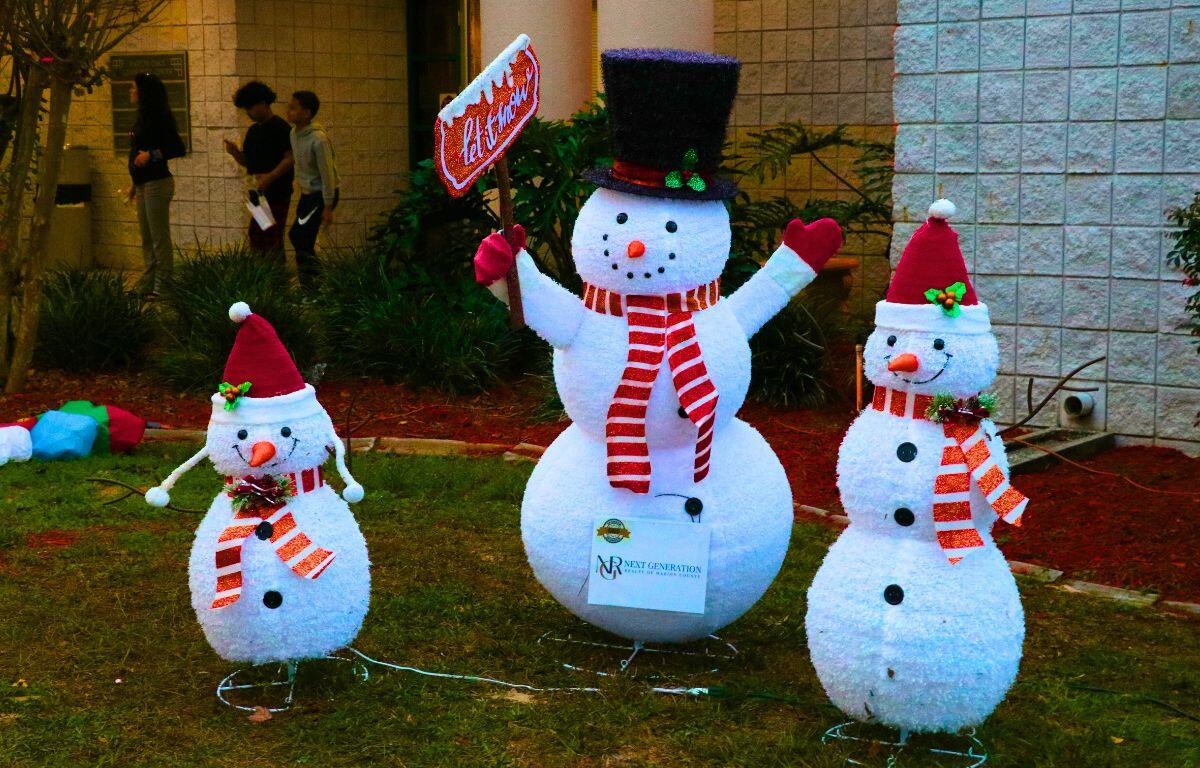 Three snowmen in a line stand outside a brick building in winter clothes.