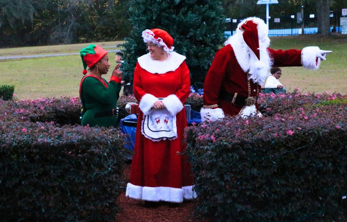 An elf, Mrs. Claus and Santa all stand in a circular bush garden with a tree in the center.