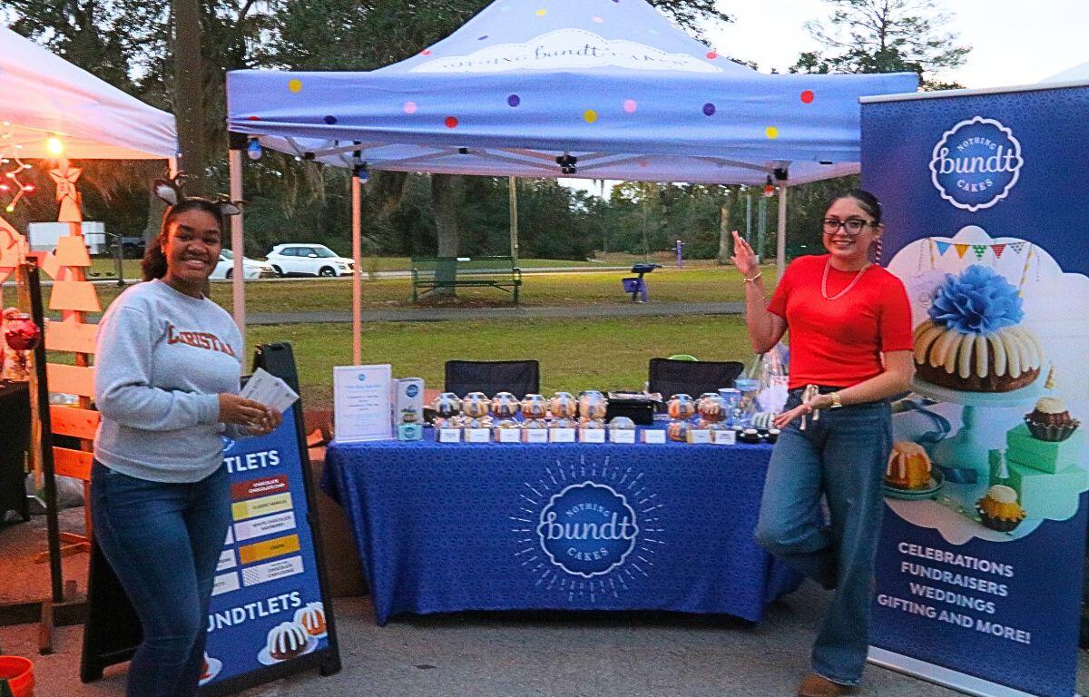 Two women pose on either side of a booth with advertising reading, "Nothing Bundt Cakes." Displayed on the table are multiple bundt cakes available for purchase.