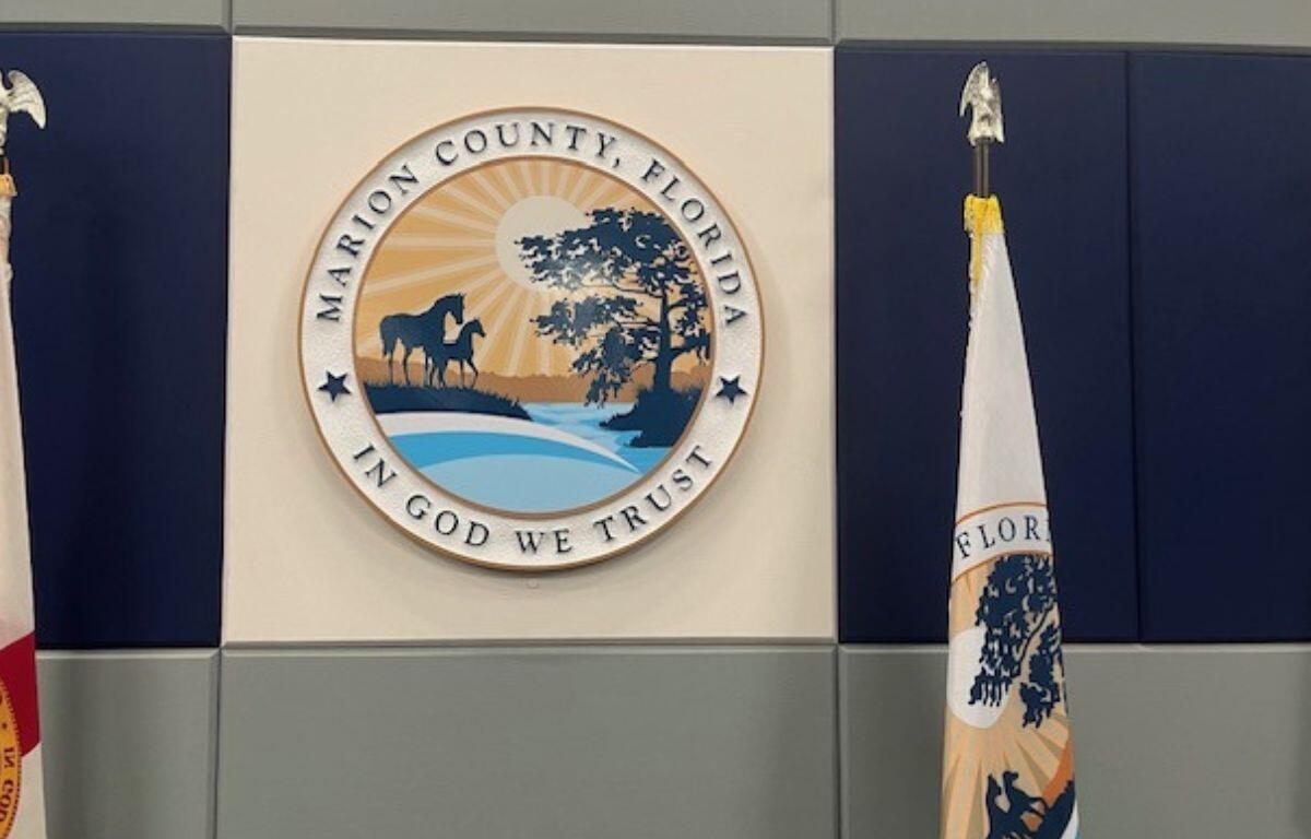 A beige background, with blue to the left and right, with flag on either side of the center beige background. A multi-colored insignia with writing an images in the center, in front of the beige background, with a gray wall underneath the insignia at the bottom of the background.