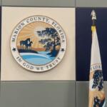 A beige background, with blue to the left and right, with flag on either side of the center beige background. A multi-colored insignia with writing an images in the center, in front of the beige background, with a gray wall underneath the insignia at the bottom of the background.