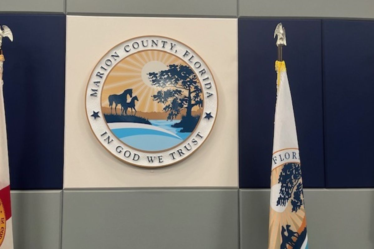 A beige background, with blue to the left and right, with flag on either side of the center beige background. A multi-colored insignia with writing an images in the center, in front of the beige background, with a gray wall underneath the insignia at the bottom of the background.