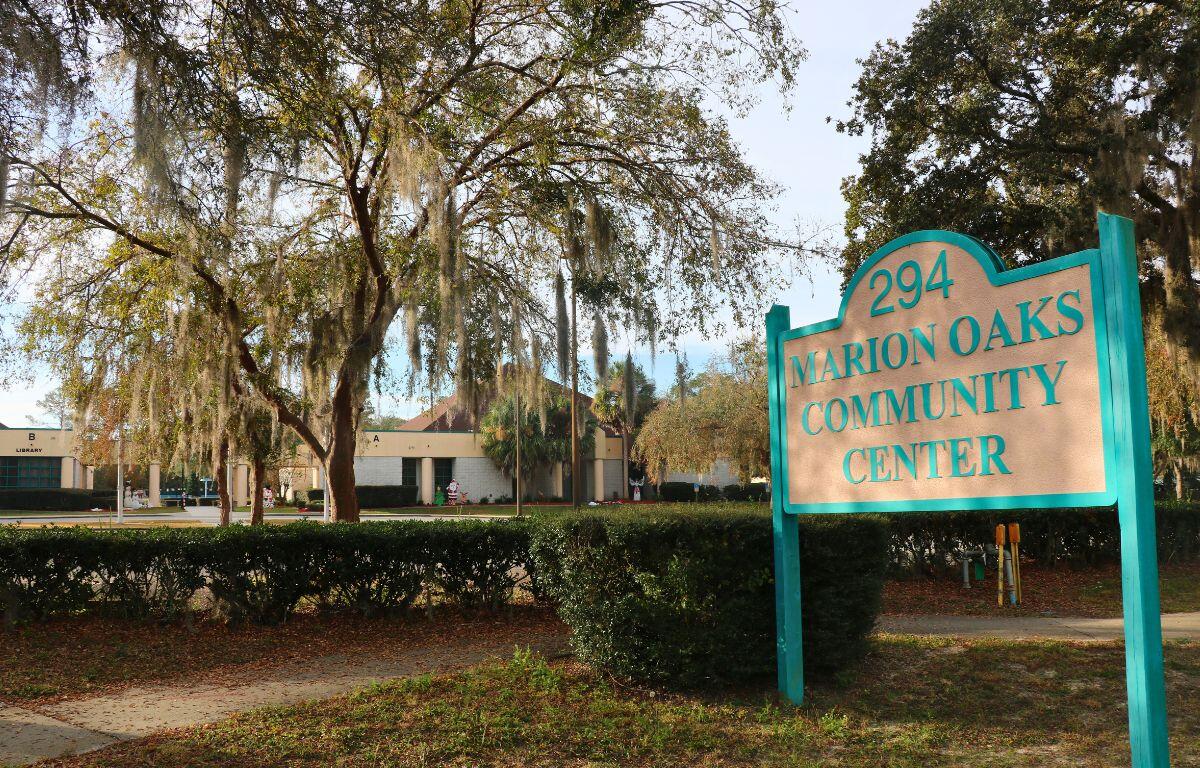 A white sign with blue accents reads, "294 Marion Oaks Community Center." In the background, bushes create a line separating a sidewalk and the community Center. Christmas decorations can be seen in front of the building.