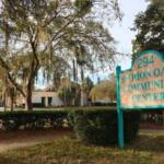 A white sign with blue accents reads, "294 Marion Oaks Community Center." In the background, bushes create a line separating a sidewalk and the community Center. Christmas decorations can be seen in front of the building.