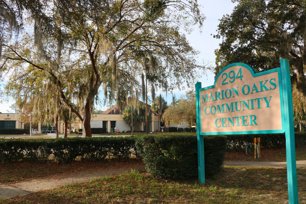 A white sign with blue accents reads, "294 Marion Oaks Community Center." In the background, bushes create a line separating a sidewalk and the community Center. Christmas decorations can be seen in front of the building.
