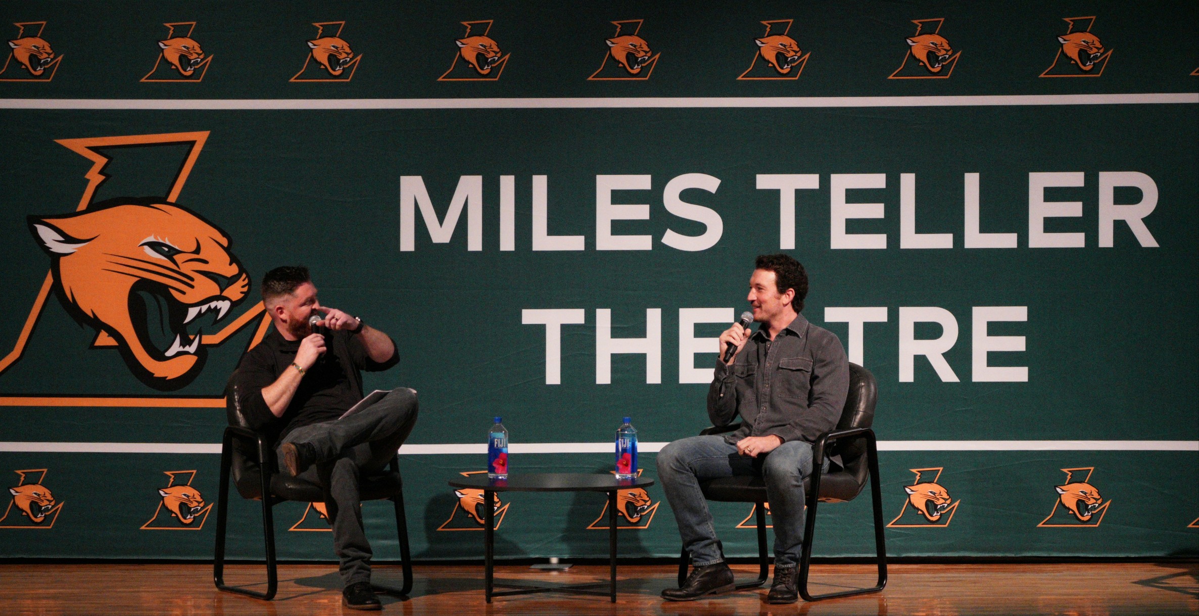Miles Teller (right) and Ryan Downs (left) sit on black chairs in front of a sign that says "Miles Teller Theatre" for a Q&A session at a special assembly on Dec 5, 2025
