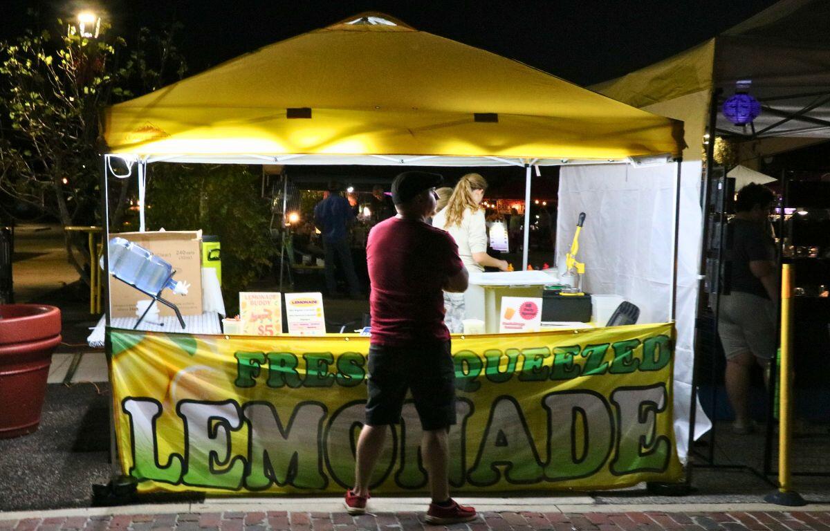 A dimly lit lemonade stand is on a paved walkway with a guest waiting for a drink.