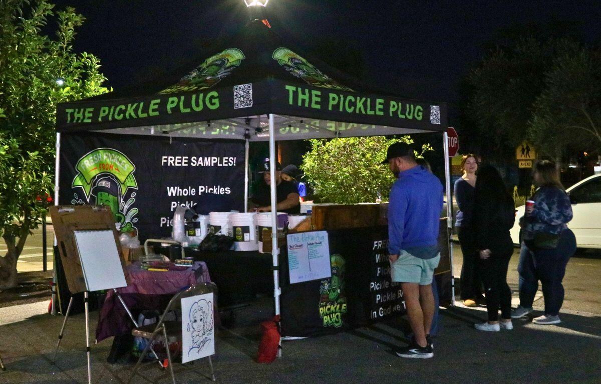 A black canopy with green writing reads, "The Pickle Plug" has guests lined up to purchase pickles.