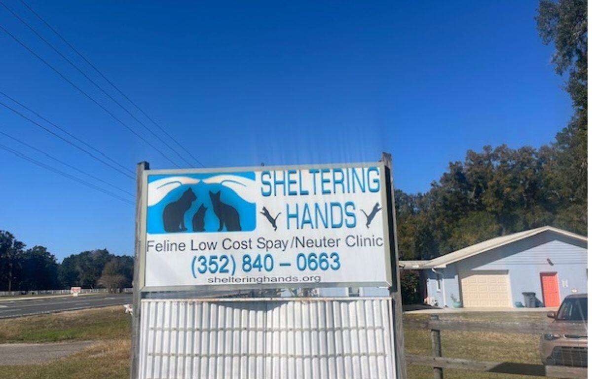 Blue sky in the background, roadway to the left, grass, and a house to the right. In the foreground a sign with lettering and images.