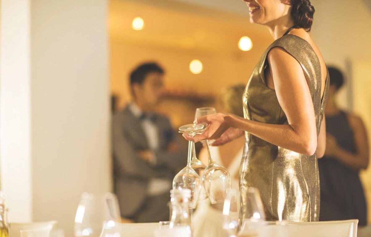 A woman in a gold dress stands in the foreground while people in suits are blurred in the background.