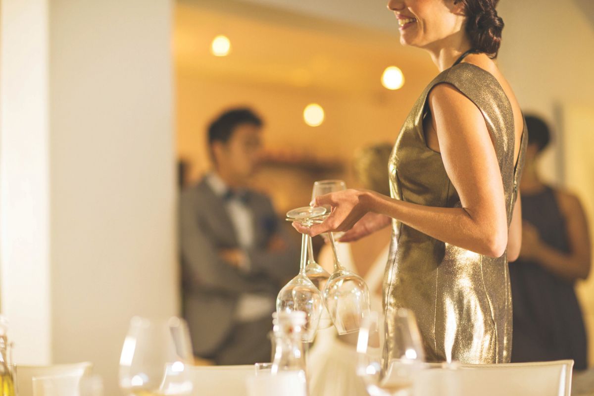 A woman in a gold dress stands in the foreground while people in suits are blurred in the background.