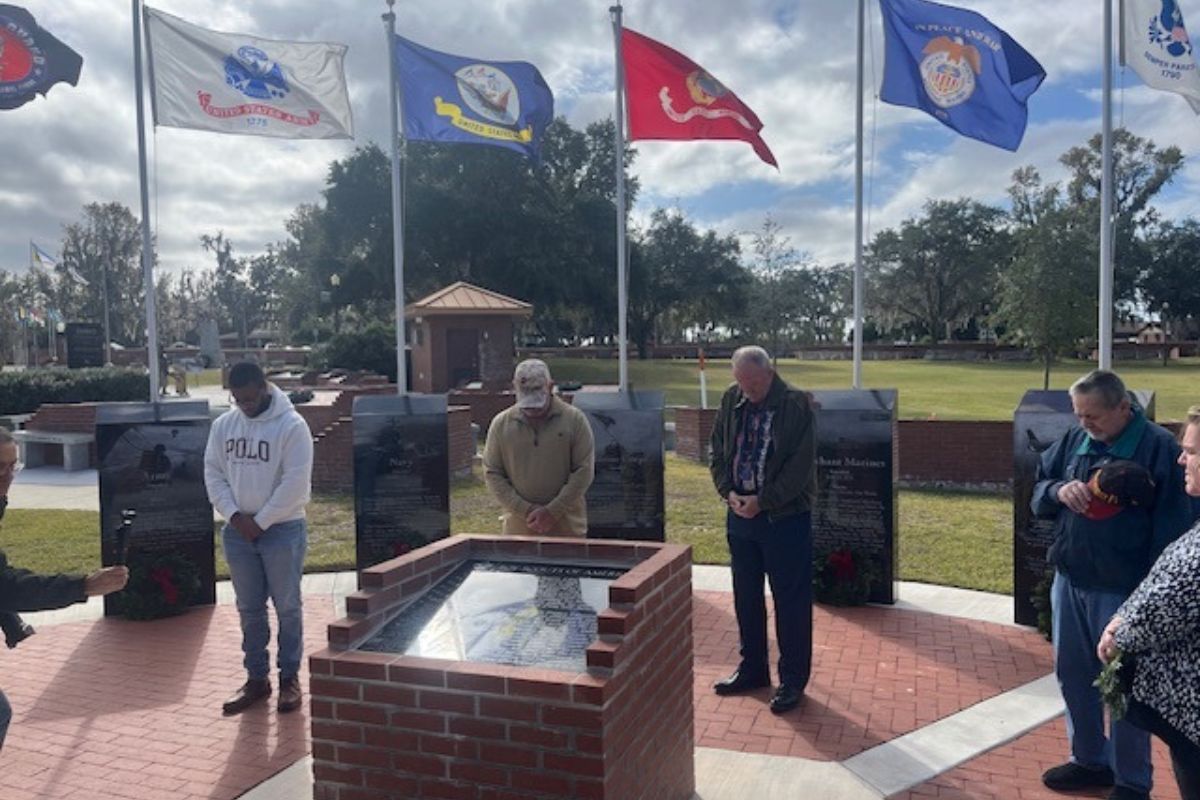 Men standing in front of flags and granite tablets with wreaths in front of the tablets, and a monument in front of the men.