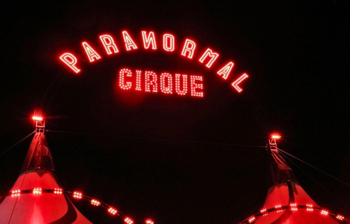 A neon sign reads, "Paranormal Cirque" above a red-lit black-and-white big top tent.