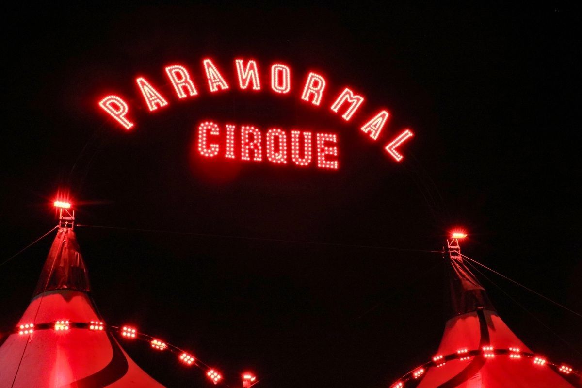 A neon sign reads, "Paranormal Cirque" above a red-lit black-and-white big top tent.
