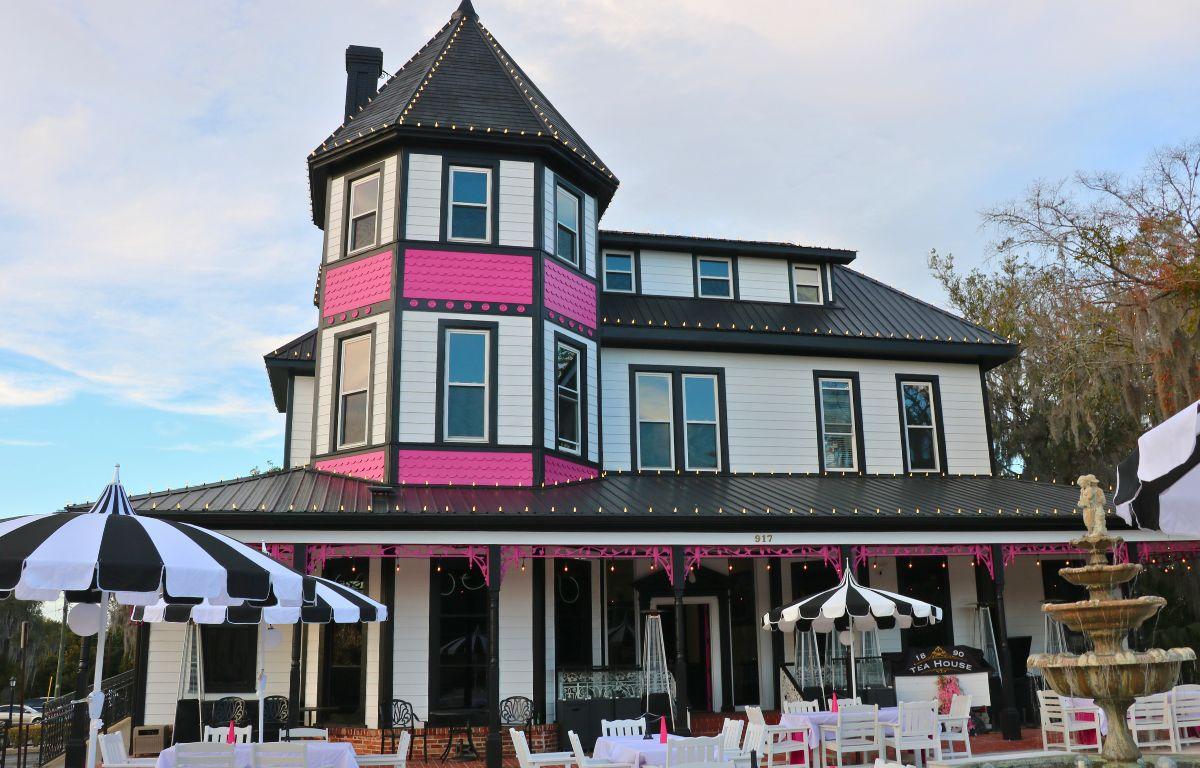 A large tea house is white with black and pink accents and lights around it. In front of the house is a patio with a fountain, tables and umbrellas providing shade on the tables.
