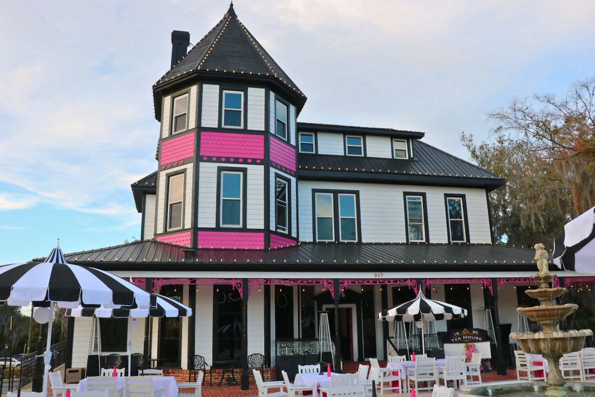 A large tea house is white with black and pink accents and lights around it. In front of the house is a patio with a fountain, tables and umbrellas providing shade on the tables.