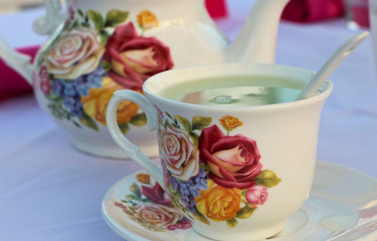A white teacup, tea plate and tea pot withe flower designs on them sit on a table with white tablecloth. In the teacup is a spoon and matcha tea.