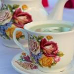 A white teacup, tea plate and tea pot withe flower designs on them sit on a table with white tablecloth. In the teacup is a spoon and matcha tea.