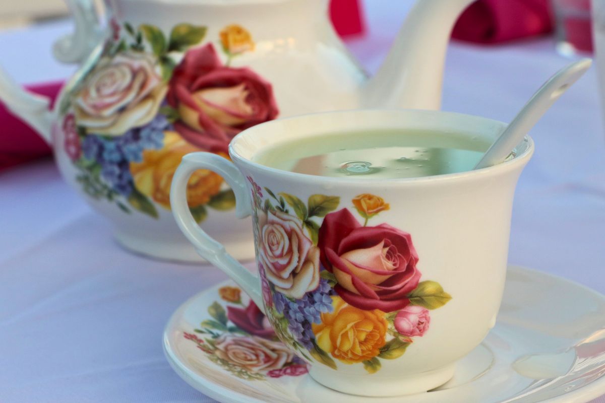 A white teacup, tea plate and tea pot withe flower designs on them sit on a table with white tablecloth. In the teacup is a spoon and matcha tea.