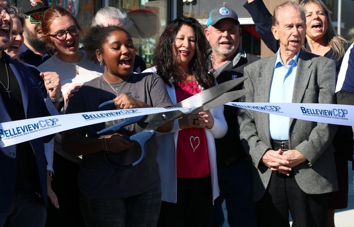 A crowd of people stand together, cheering as a young woman cuts a white ribbon with blue letters reading, "Belleview CEP."