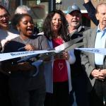 A crowd of people stand together, cheering as a young woman cuts a white ribbon with blue letters reading, "Belleview CEP."