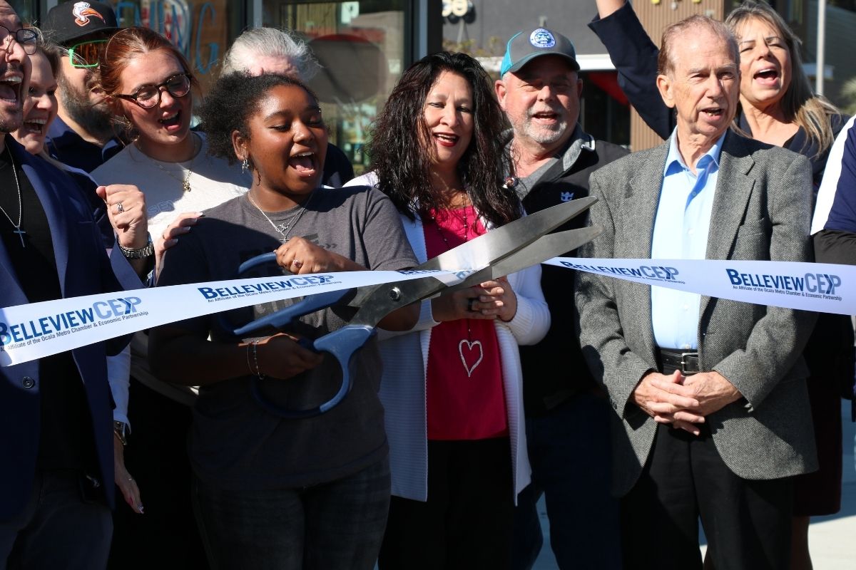 A crowd of people stand together, cheering as a young woman cuts a white ribbon with blue letters reading, "Belleview CEP."