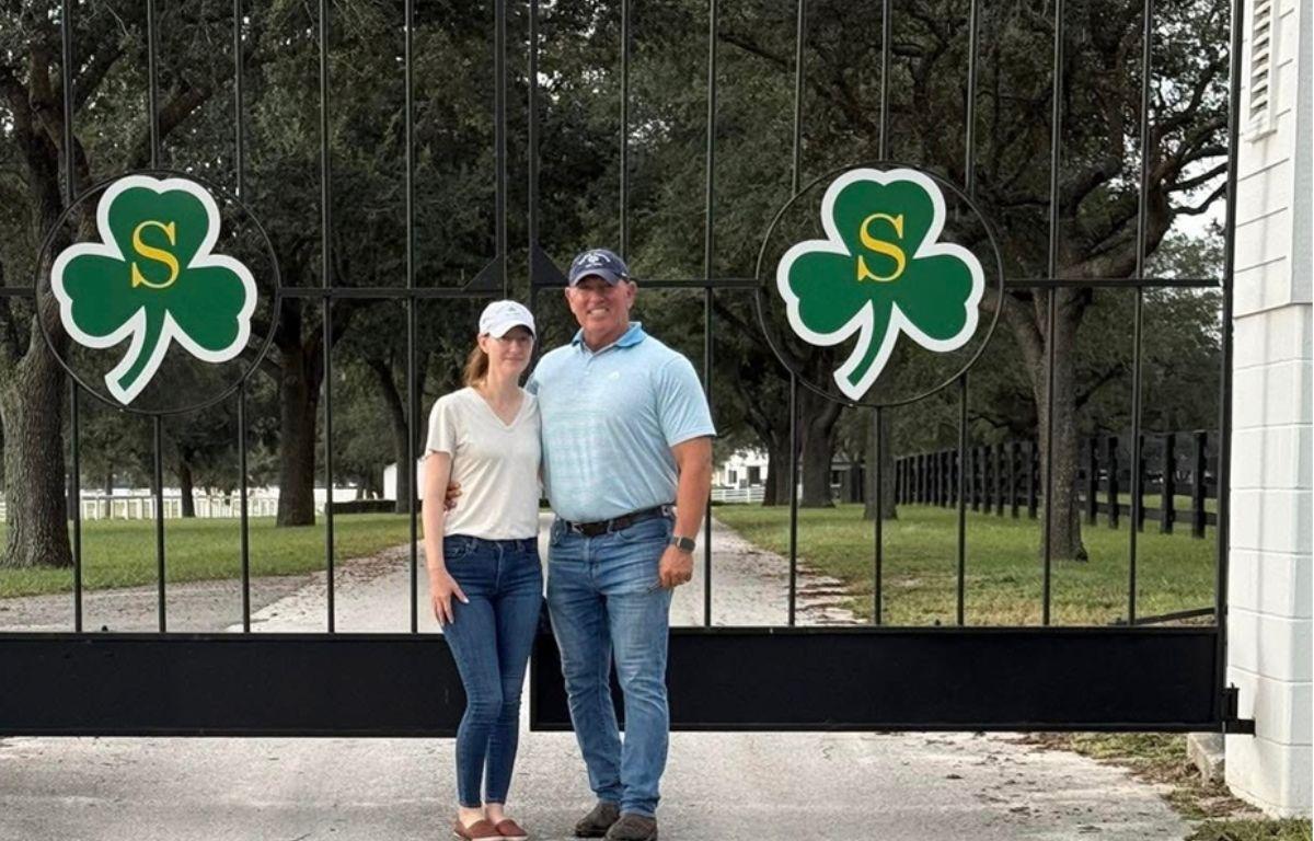 A man and a woman standing on pavement in front of an iron gate with shamrock emblems on the front of the gate, with grass and trees in the background.