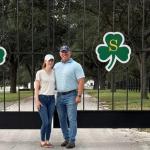 A man and a woman standing on pavement in front of an iron gate with shamrock emblems on the front of the gate, with grass and trees in the background.
