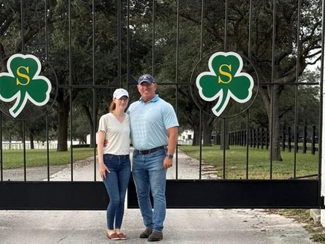 A man and a woman standing on pavement in front of an iron gate with shamrock emblems on the front of the gate, with grass and trees in the background.