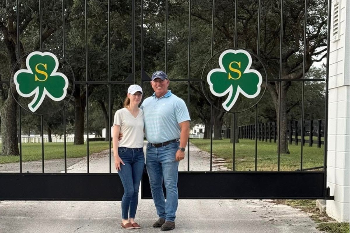 A man and a woman standing on pavement in front of an iron gate with shamrock emblems on the front of the gate, with grass and trees in the background.