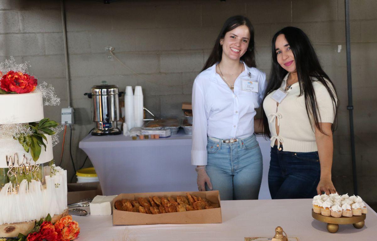 Two women stand behind a booth with a decorative tiered cake with flowers on it to the left, empanadas in the center and small cupcakes to the right.