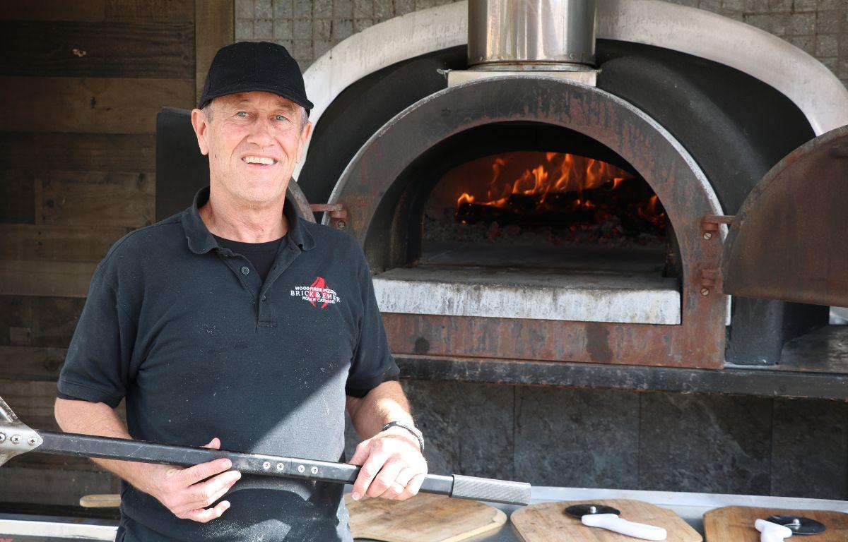 A man stands in front of an open metal oven with wood board and pizza cutters in the background.