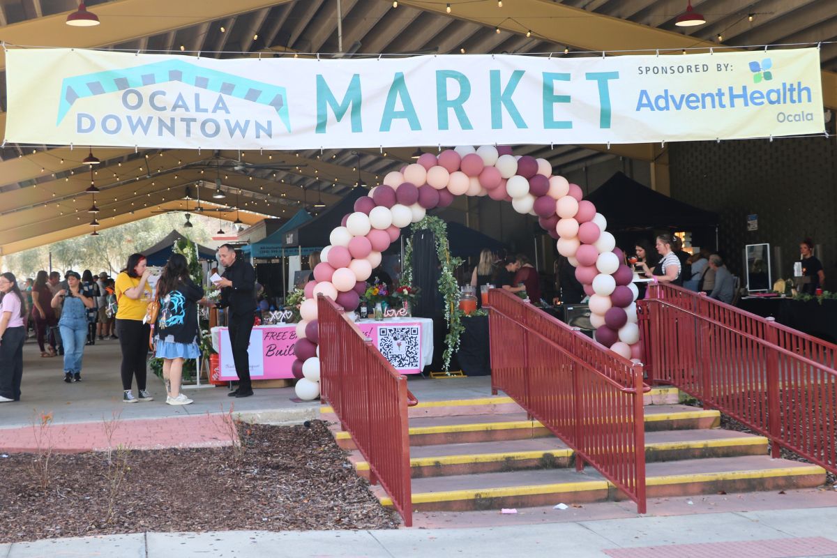 A large, open-air event area features a large balloon arch at the entrance with a white banner that reads, "Ocala Downtown Market. Sponsored by: Advent Health." A large market is taking place behind the entrance.