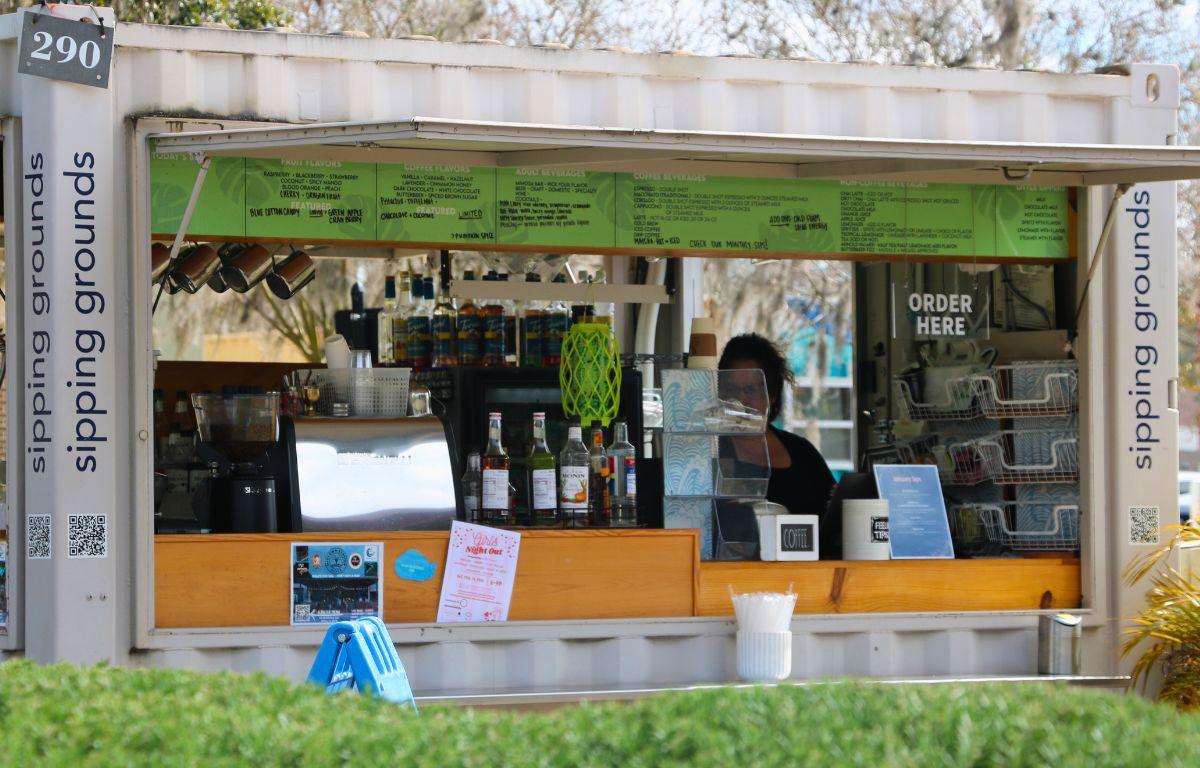A coffee stand features menus and a straw cup around the stand. On the sides of the building, black words read, "Sipping Grounds."