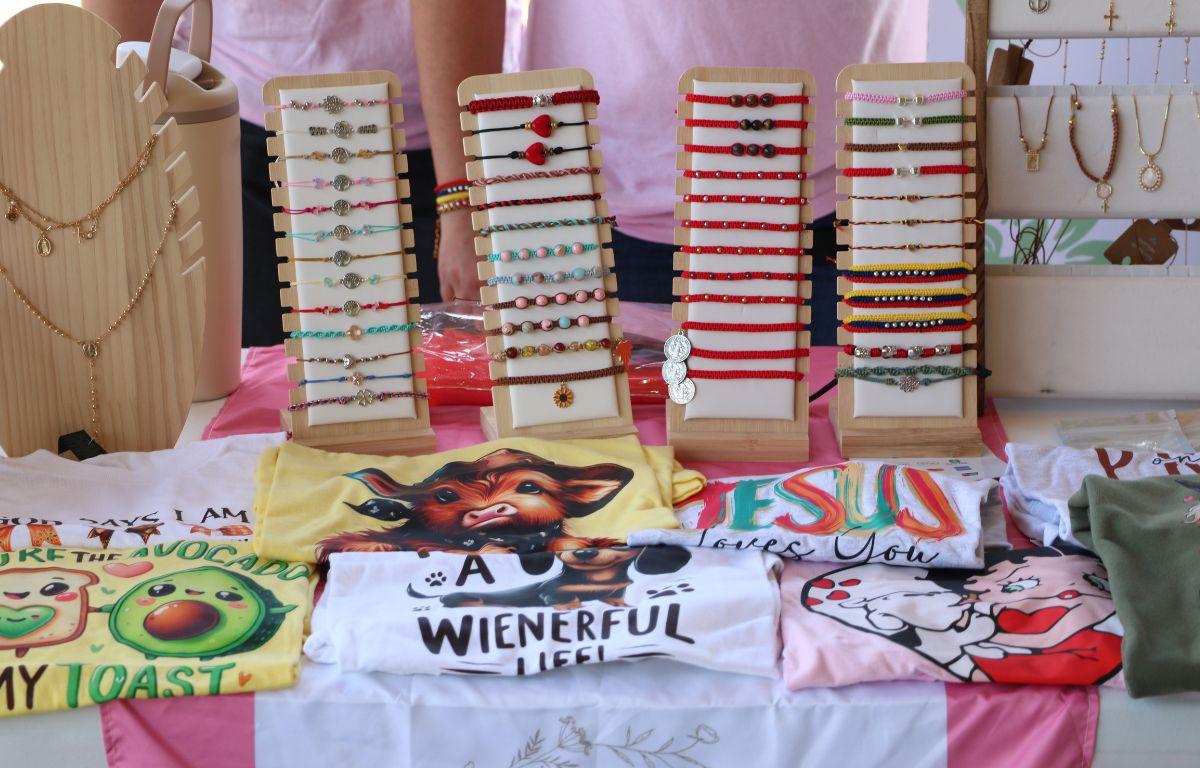 A table with a pink and white tablecloth features jewelry and shirts.