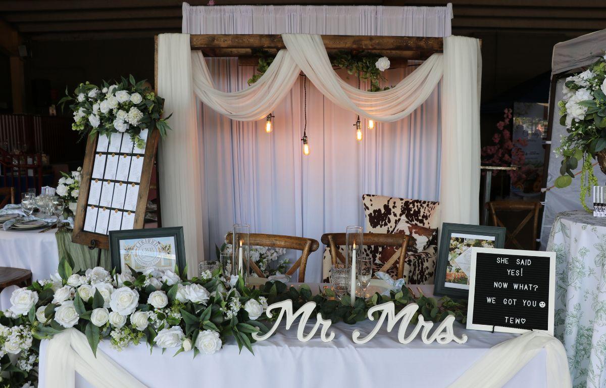 A booth at a market features a picturesque background with flowing drapes, flowers, chairs, candles and a sign with white letters on it. The sign reads, "She said yes! Now what? We got you tew."