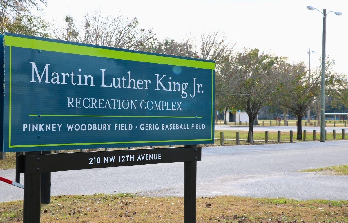 A blue sign with green accents and brown metal supports features white text reading, "Martin Luther King Jr. Recreation Complex. Pinkney Woodbury Field · Gerig Baseball Field. 210 NW 12th Avenue." The sign stands at the edge of a recreation park complex.