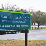 A blue sign with green accents and brown metal supports features white text reading, "Martin Luther King Jr. Recreation Complex. Pinkney Woodbury Field · Gerig Baseball Field. 210 NW 12th Avenue." The sign stands at the edge of a recreation park complex.
