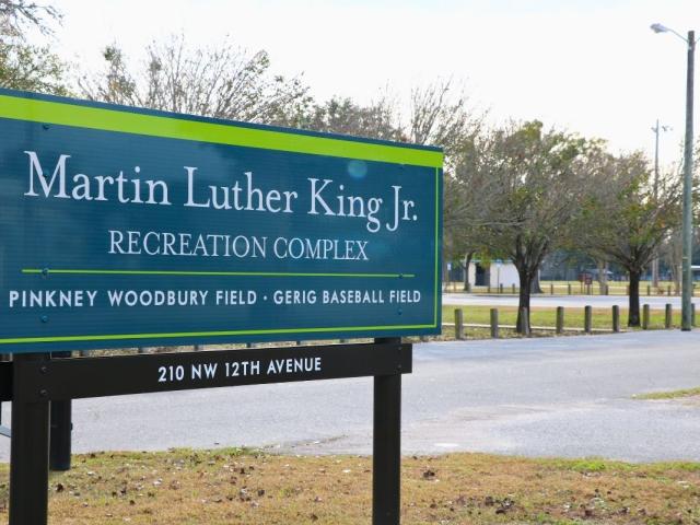 A blue sign with green accents and brown metal supports features white text reading, "Martin Luther King Jr. Recreation Complex. Pinkney Woodbury Field · Gerig Baseball Field. 210 NW 12th Avenue." The sign stands at the edge of a recreation park complex.