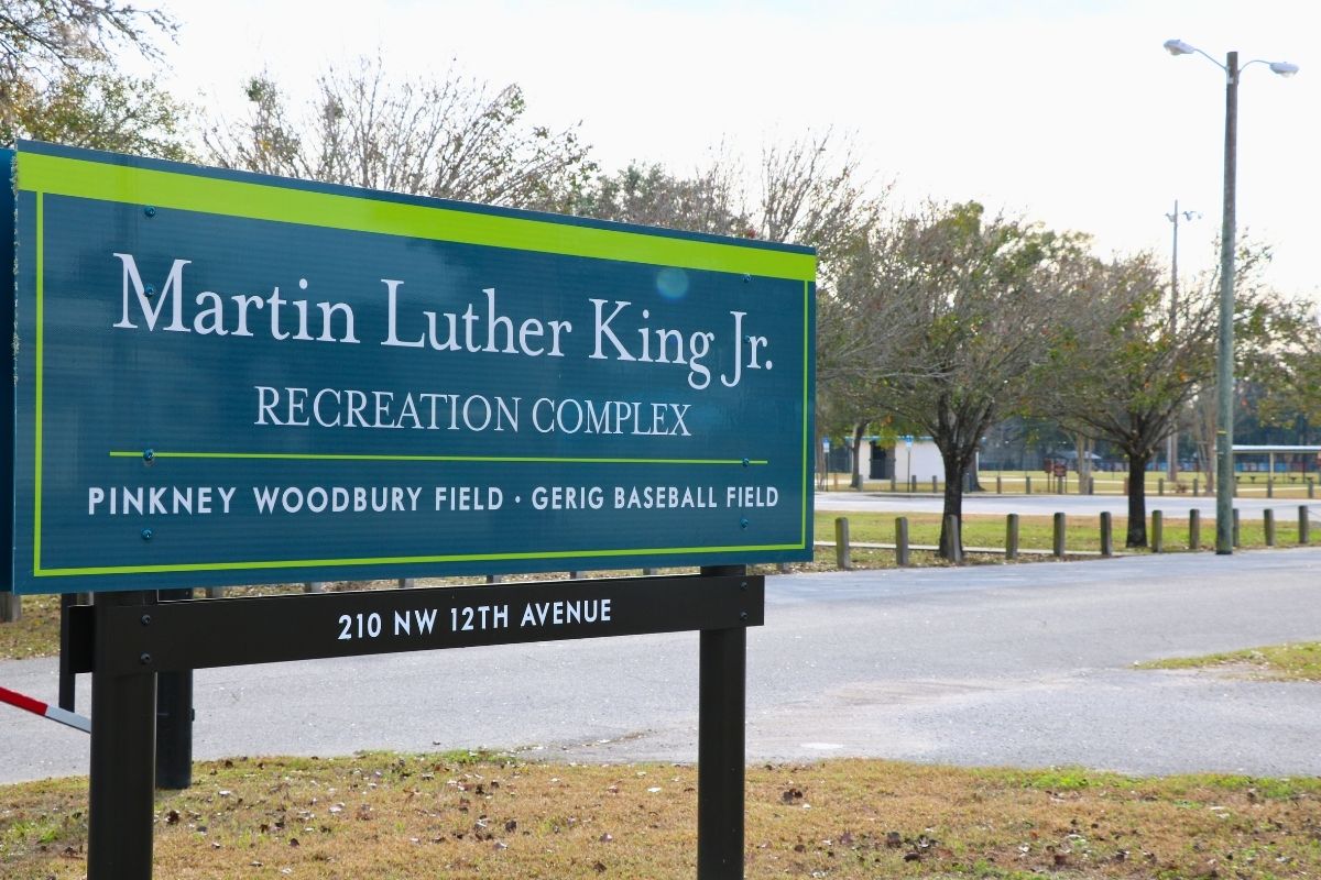 A blue sign with green accents and brown metal supports features white text reading, "Martin Luther King Jr. Recreation Complex. Pinkney Woodbury Field · Gerig Baseball Field. 210 NW 12th Avenue." The sign stands at the edge of a recreation park complex.