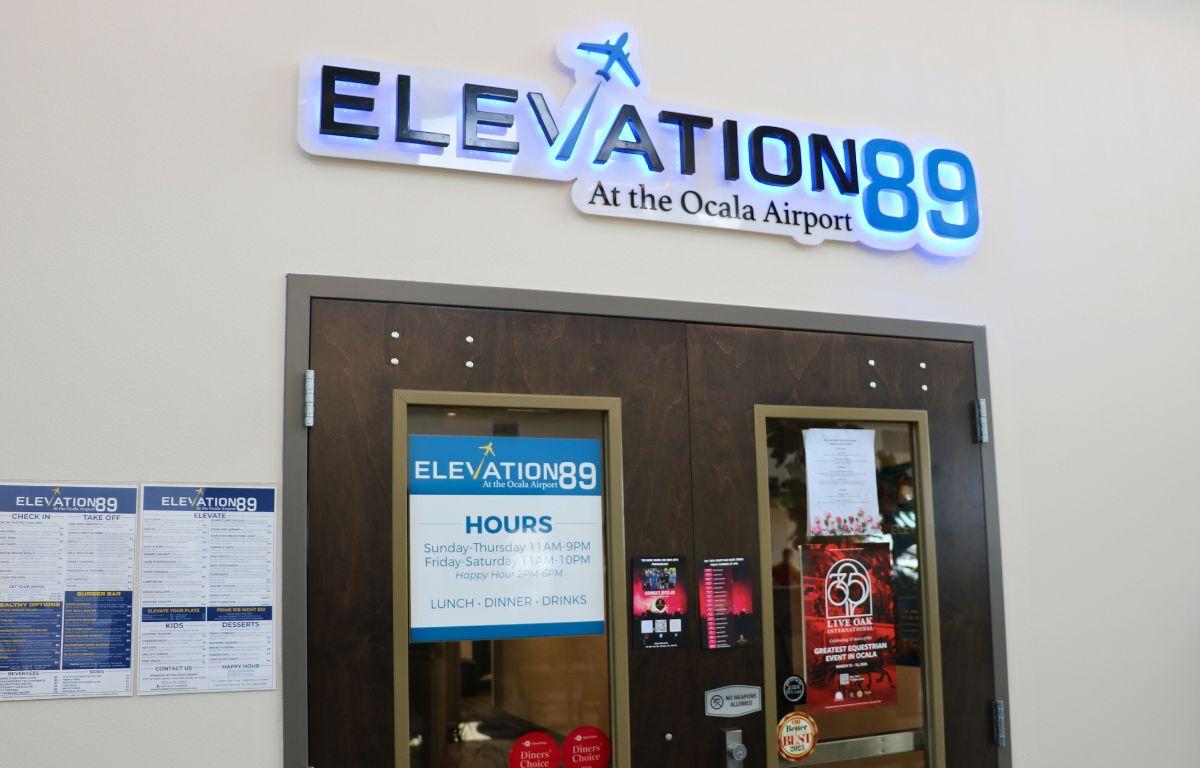 The entrance of a restaurant has a light-up sign that reads, "Elevation 89 at the airport." Underneath the sign is a wood door with posters strewn on it.