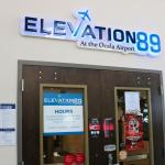 The entrance of a restaurant has a light-up sign that reads, "Elevation 89 at the airport." Underneath the sign is a wood door with posters strewn on it.