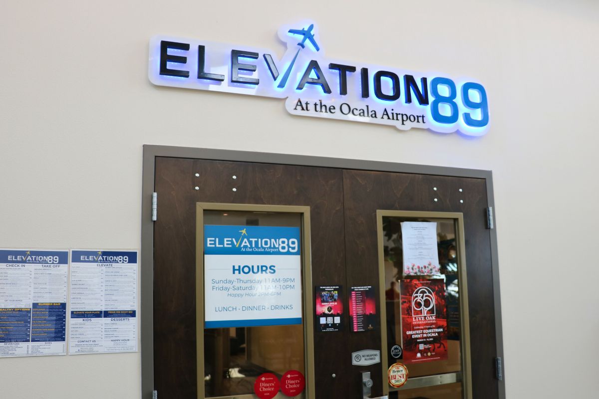 The entrance of a restaurant has a light-up sign that reads, "Elevation 89 at the airport." Underneath the sign is a wood door with posters strewn on it.